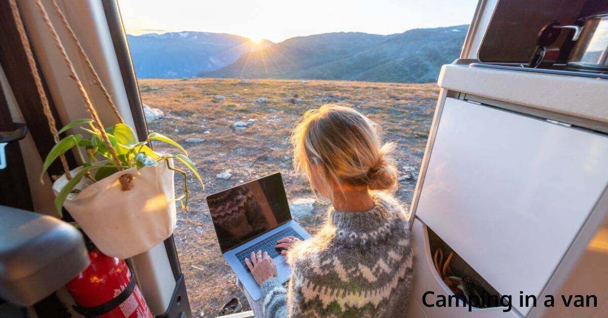 woman seated at camping van door using a laptop with sunrise in the background